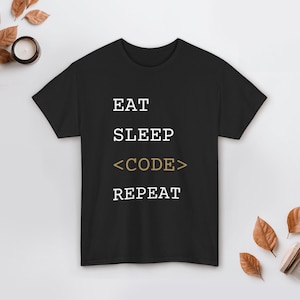 Può includere: T-shirt nero con le parole "EAT SLEEP <CODE> REPEAT" stampate in bianco e oro. La maglietta è girocollo e ha maniche corte. La maglietta è realizzata in un materiale morbido.