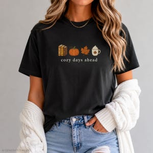 Op de afbeelding: Een zwart t-shirt met korte mouwen en een afbeelding van drie boeken, een oranje pompoen, een bruin esdoornblad en een witte mok met slagroom. Onder de iconen staat de tekst "cozy days ahead" in een eenvoudig wit schreeflettertype.