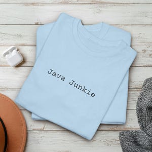 Op de afbeelding: Twee lichtblauwe T-shirts liggen op een licht houten oppervlak. Op het bovenste shirt staat in zwarte letters "Java Junkie". Een paar witte draadloze oortelefoons en een bruine hoed staan op de achtergrond.