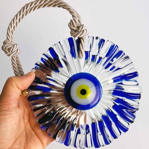 Op de afbeelding: Een blauwe en witte glazen amulet met een touwlus om op te hangen. Het boze oog is omgeven door een helder glasontwerp met blauwe accenten.