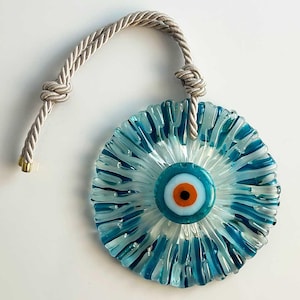 Op de afbeelding: Een blauwe en witte glazen boze oog charme met een witte touw hanger. Het boze oog is omgeven door een blauw en wit swirl patroon.