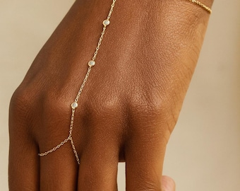 May include: A delicate gold hand chain bracelet. The bracelet features a fine chain that connects to a ring and a bracelet chain. It is adorned with small, clear gemstones. The jewelry is displayed on a hand.
