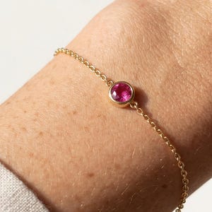 Peut inclure: Un bracelet en or fin avec une seule pierre précieuse rose vif et ronde. Le bracelet est composé d'une fine chaîne, avec la pierre précieuse sertie dans une lunette simple. Le bijou est porté au poignet, soulignant l'élégance simple de la pièce.