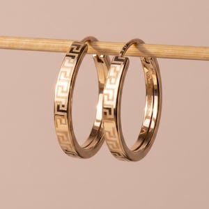 Könnte beinhalten: Ein Paar goldfarbene Creolen mit griechischem Schlüsselmuster. Die Ohrringe hängen an einem Holzstab vor einem sanften rosa Hintergrund. Die Ohrringe sind rund und haben ein klassisches Design.
