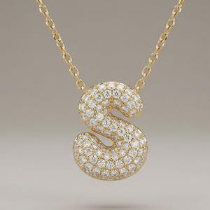 May include: A gold necklace with a pendant shaped like the letter 'S'. The pendant is covered in small, sparkling diamonds. The chain is also gold and fine. The necklace is a piece of jewellery.