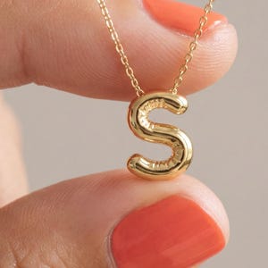 Op de afbeelding: Een goudkleurige ketting met een hanger in de vorm van de letter "S". De hanger heeft een afgerond, bubbelachtig ontwerp. De ketting wordt vastgehouden door een persoon met oranje nagellak.