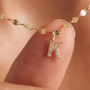 May include: A delicate gold necklace featuring a small, diamond-studded letter "K" pendant. The chain is made of small, flat, circular links. The necklace is displayed against a blurred background.