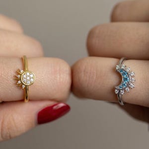 May include: Two rings, one gold and one silver, are displayed. The gold ring features a sunburst design with a central cluster of small, clear stones. The silver ring showcases a crescent moon shape with blue stones and surrounding accents. Jewelry.