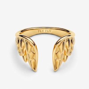 May include: A gold-toned open ring featuring a stylized wing design on both sides. The band is polished and smooth, with the text FLY FLY engraved clearly on the inner surface of the ring. The design is symmetrical, showcasing detailed feather textures.