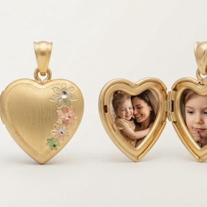 Può includere: Ciondolo a forma di cuore dorato con dettagli floreali. Un medaglione è chiuso, l'altro è aperto, rivelando due foto. Il medaglione è un ciondolo, progettato per custodire ricordi. È un bellissimo gioiello.