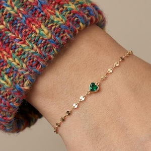 Puede incluir: Un brazalete dorado con una pequeña gema verde en forma de corazón. El brazalete está en una muñeca, con una manga de suéter de punto colorido en el fondo. La cadena está hecha de pequeños eslabones circulares planos.