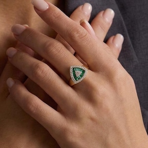 Könnte beinhalten: Ein goldener Ring mit einem dreieckigen Smaragd-Mittelstein, umgeben von kleinen weißen Edelsteinen. Der Ring wird auf einer Hand vor einem dunkelgrauen Blazer gezeigt. Das Design des Rings ist geometrisch und elegant.