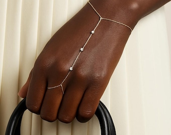 May include: A delicate gold hand chain bracelet with three small, sparkling gemstones. The bracelet connects to a ring and wrist chain. The hand is holding a black handbag, set against a cream-colored background.