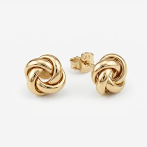 May include: A pair of gold-colored stud earrings. Each earring features a knot design, with a polished, reflective surface. The earrings are set against a white background, highlighting their intricate form and warm hue.