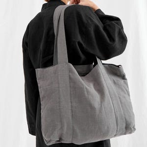 May include: A gray tote bag with long straps is carried over the shoulder. The bag is made of a textured fabric and has a rectangular shape. The person in the image is wearing a black top.