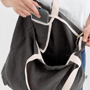 May include: Dark gray tote bag with cream-colored straps and trim. The bag has a large open pocket and an interior pocket holding a smartphone. The bag is made of a textured fabric.