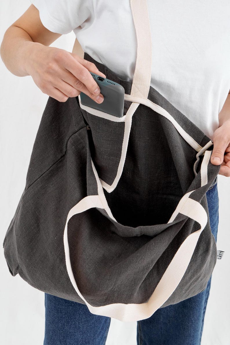 May include: Dark gray tote bag with cream-colored straps and trim. The bag has a large open pocket and an interior pocket holding a smartphone. The bag is made of a textured fabric.
