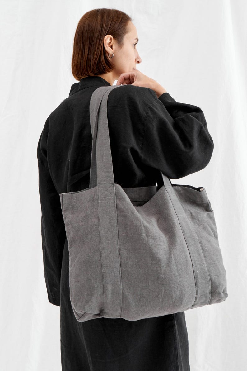 May include: A gray tote bag with long straps is carried over the shoulder. The bag is made of a textured fabric and has a rectangular shape. The person in the image is wearing a black top.