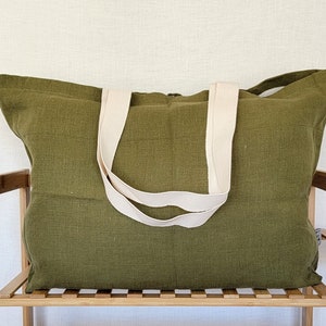 May include: Olive green tote bag with cream-colored straps. The bag is made of a textured fabric and is sitting on a wooden shelf. The tote bag is a large size, suitable for carrying various items.