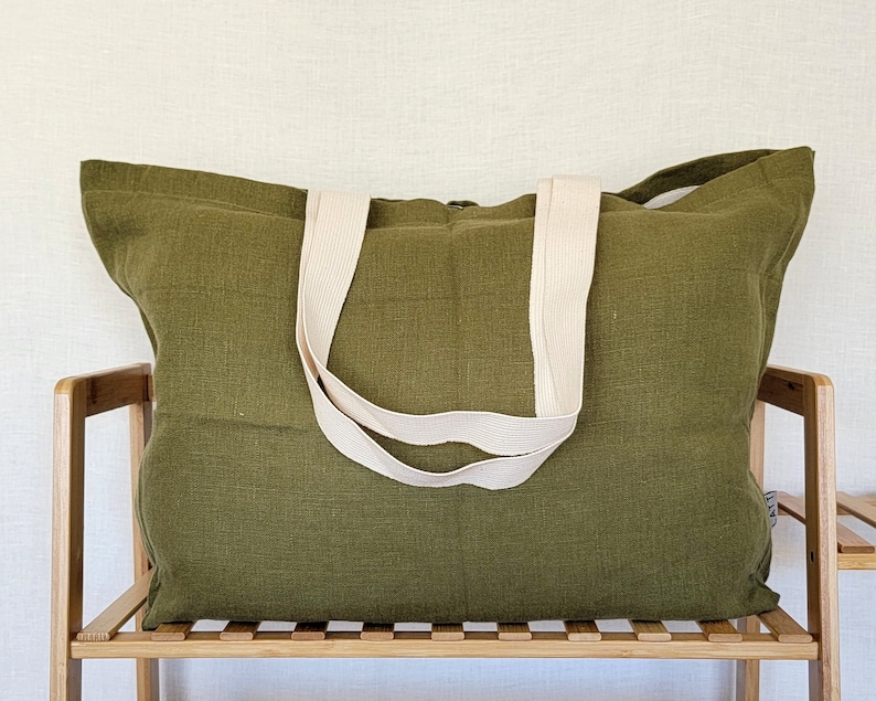 May include: Olive green tote bag with cream-colored straps. The bag is made of a textured fabric and is sitting on a wooden shelf. The tote bag is a large size, suitable for carrying various items.