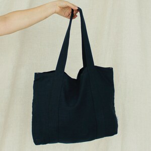 May include: A navy blue tote bag with long handles, held up by a person. The bag is made of a textured fabric and has a simple, minimalist design. The bag is a solid color with no visible patterns or embellishments.
