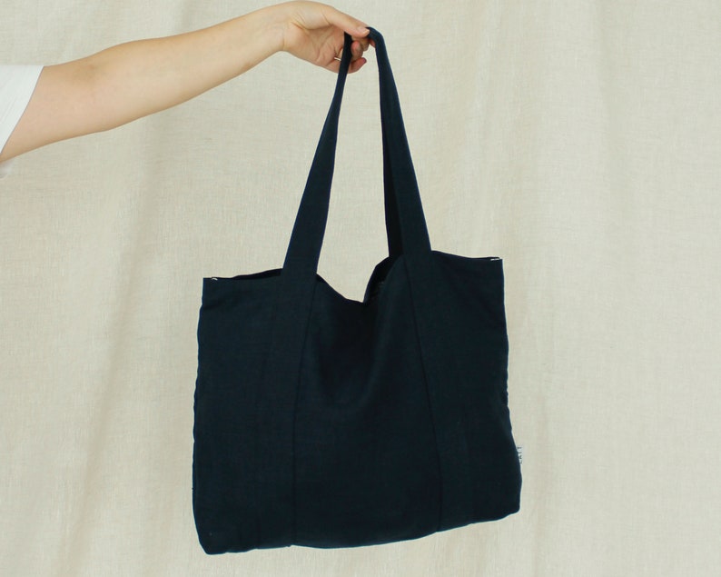 May include: A navy blue tote bag with long handles, held up by a person. The bag is made of a textured fabric and has a simple, minimalist design. The bag is a solid color with no visible patterns or embellishments.