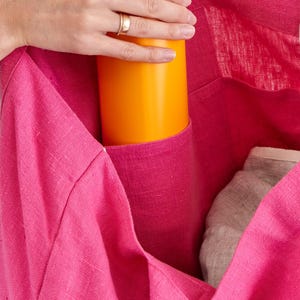 May include: A bright pink tote bag with a pocket holding an orange bottle and a gray item. The bag is made of a textured fabric. The top of the bottle is pink.