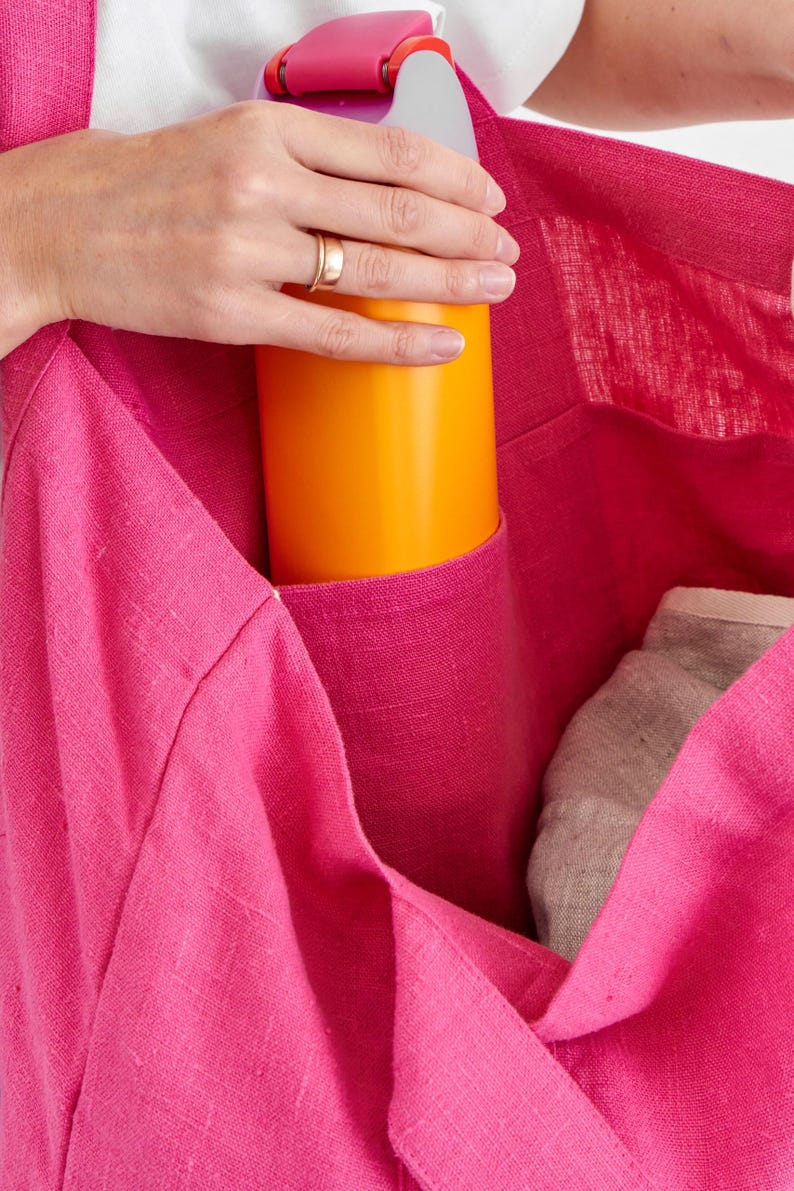 May include: A bright pink tote bag with a pocket holding an orange bottle and a gray item. The bag is made of a textured fabric. The top of the bottle is pink.