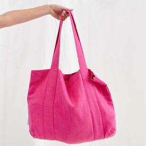 May include: A vibrant pink tote bag with long handles. The bag is made of a textured fabric and has a simple, rectangular shape. The bag is held up by a hand against a white background. The bag is a bright, solid color.