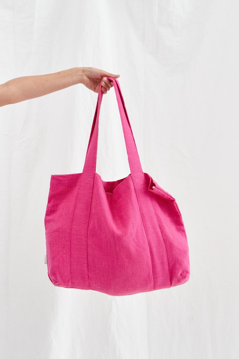 May include: A vibrant pink tote bag with long handles. The bag is made of a textured fabric and has a simple, rectangular shape. The bag is held up by a hand against a white background. The bag is a bright, solid color.