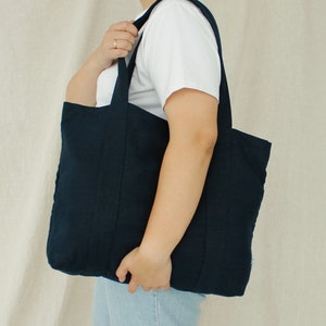 May include: A navy blue tote bag with two handles is held by a person. The bag is made of a textured fabric and has a rectangular shape. The person is wearing a white t-shirt and blue jeans.