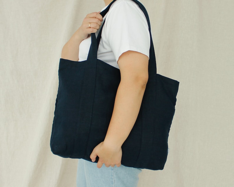 May include: A navy blue tote bag with two handles is held by a person. The bag is made of a textured fabric and has a rectangular shape. The person is wearing a white t-shirt and blue jeans.