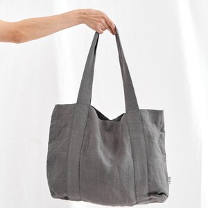 May include: A gray tote bag with long handles, held up against a white background. The bag is made of a textured fabric and has a rectangular shape. The handles are sewn to the bag's body.