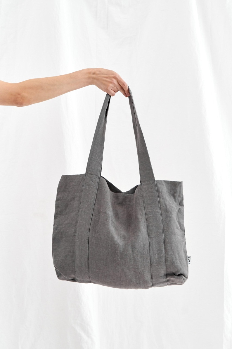 May include: A gray tote bag with long handles, held up against a white background. The bag is made of a textured fabric and has a rectangular shape. The handles are sewn to the bag's body.