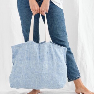 May include: A light blue linen tote bag with white handles. The bag is held by a person wearing blue jeans. The tote bag is a soft, casual style, suitable for carrying groceries or other items.