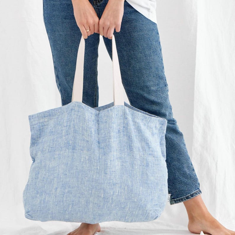 May include: A light blue linen tote bag with white handles. The bag is held by a person wearing blue jeans. The tote bag is a soft, casual style, suitable for carrying groceries or other items.