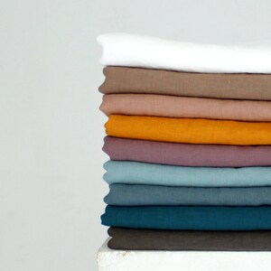 May include: A stack of folded linen fabric in various colors, including white, brown, orange, purple, and teal. The fabrics are neatly arranged, showcasing their different hues and textures. The image is set against a plain white background.