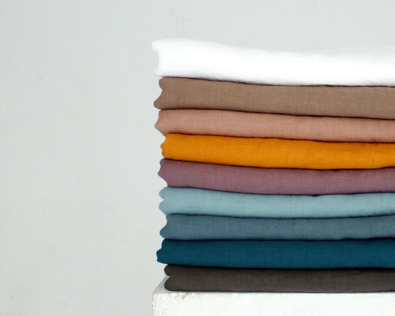 May include: A stack of folded linen fabric in various colors, including white, brown, orange, purple, and teal. The fabrics are neatly arranged, showcasing their different hues and textures. The image is set against a plain white background.