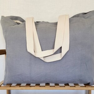 May include: A large, rectangular gray tote bag with cream-colored straps. The bag is made of a textured fabric and sits on a wooden shelf. The straps are wide and appear to be made of a sturdy material. The bag is designed for carrying items.