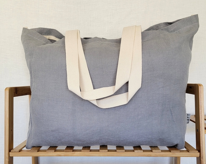 May include: A large, rectangular gray tote bag with cream-colored straps. The bag is made of a textured fabric and sits on a wooden shelf. The straps are wide and appear to be made of a sturdy material. The bag is designed for carrying items.