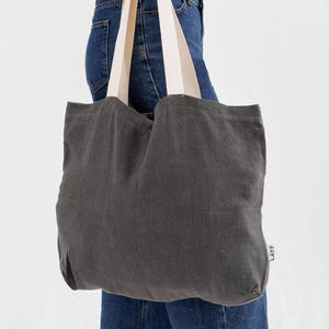 May include: A dark gray tote bag with cream-colored straps is held by a person wearing blue jeans and a white shirt. The bag is made of a textured fabric and has a simple, minimalist design. The bag has the word "LATT" printed on the side.