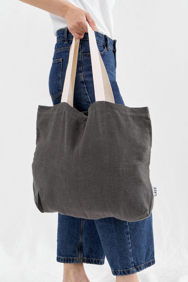 May include: A dark gray tote bag with cream-colored straps is held by a person wearing blue jeans and a white shirt. The bag is made of a textured fabric and has a simple, minimalist design. The bag has the word "LATT" printed on the side.