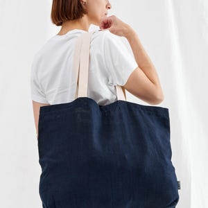 May include: A navy blue tote bag with cream-colored straps. The bag is made of a textured fabric and is being carried over the shoulder. The background is a plain white.