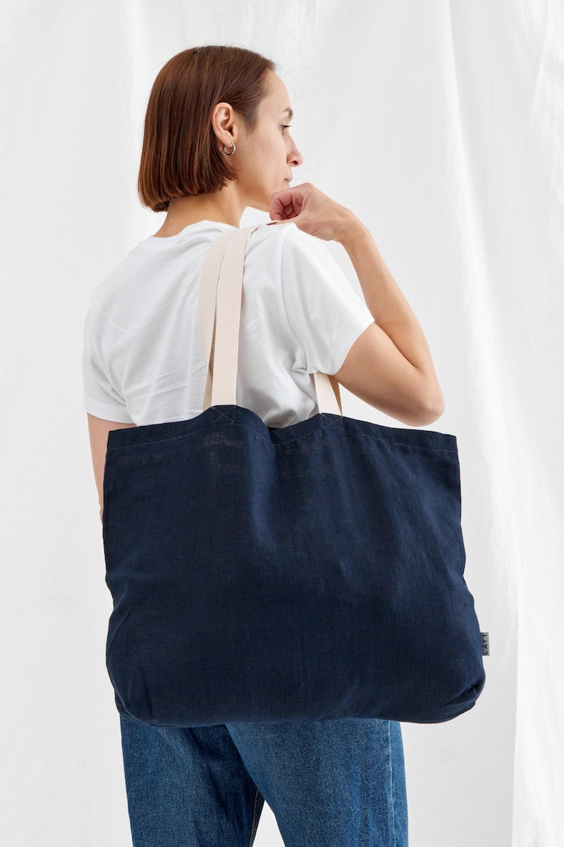 May include: A navy blue tote bag with cream-colored straps. The bag is made of a textured fabric and is being carried over the shoulder. The background is a plain white.