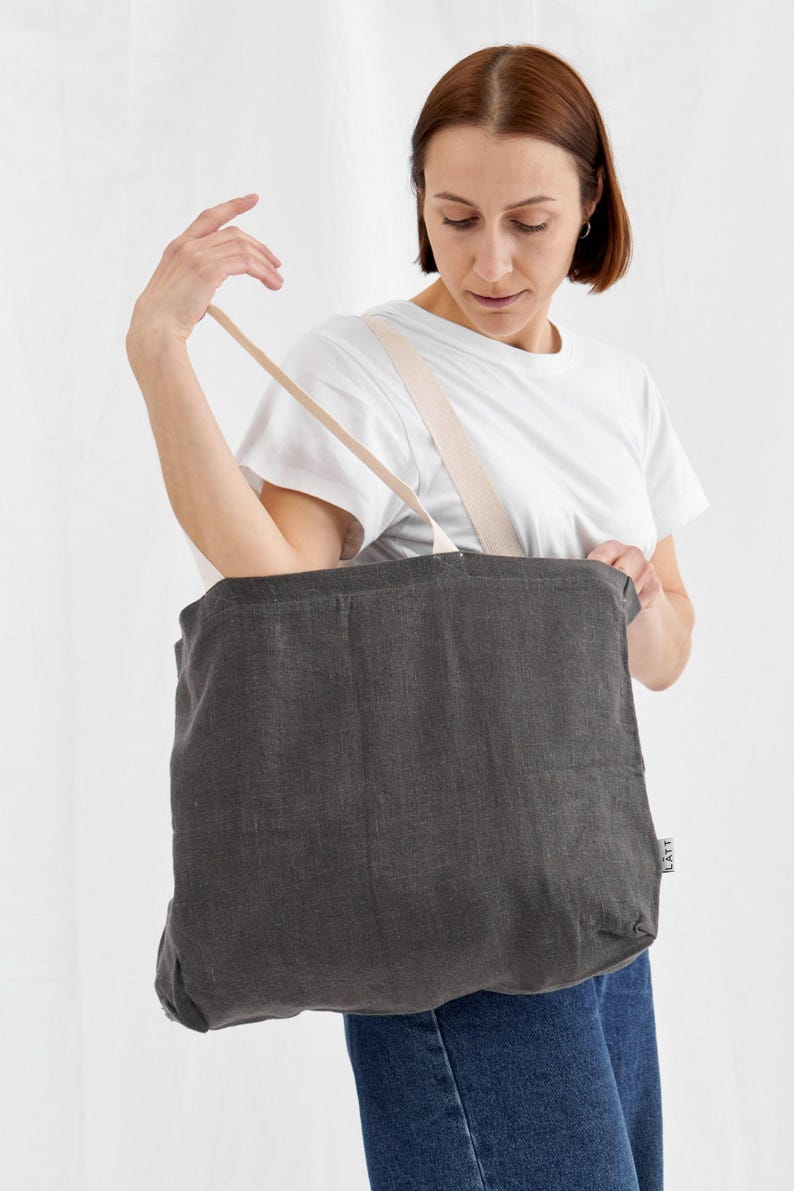 May include: A dark gray tote bag with beige straps is held up. The bag is made of a textured fabric. The person in the image is wearing a white t-shirt and blue jeans. The bag has a small tag that says "LATT".