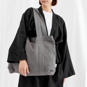May include: A gray tote bag with a rectangular shape and a long strap. The bag is made of a textured fabric and has a simple, minimalist design. The person in the image is wearing a black kimono-style jacket and a white t-shirt.