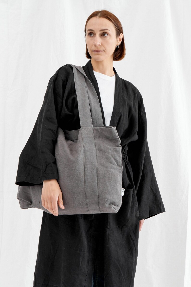 May include: A gray tote bag with a rectangular shape and a long strap. The bag is made of a textured fabric and has a simple, minimalist design. The person in the image is wearing a black kimono-style jacket and a white t-shirt.