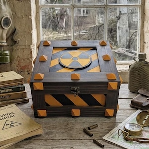 May include: A wooden box with a radiation symbol and orange and black hazard stripes. The box is surrounded by survival gear, including a gas mask, books, a compass, and a water bottle. The scene evokes a post-apocalyptic or survivalist theme.