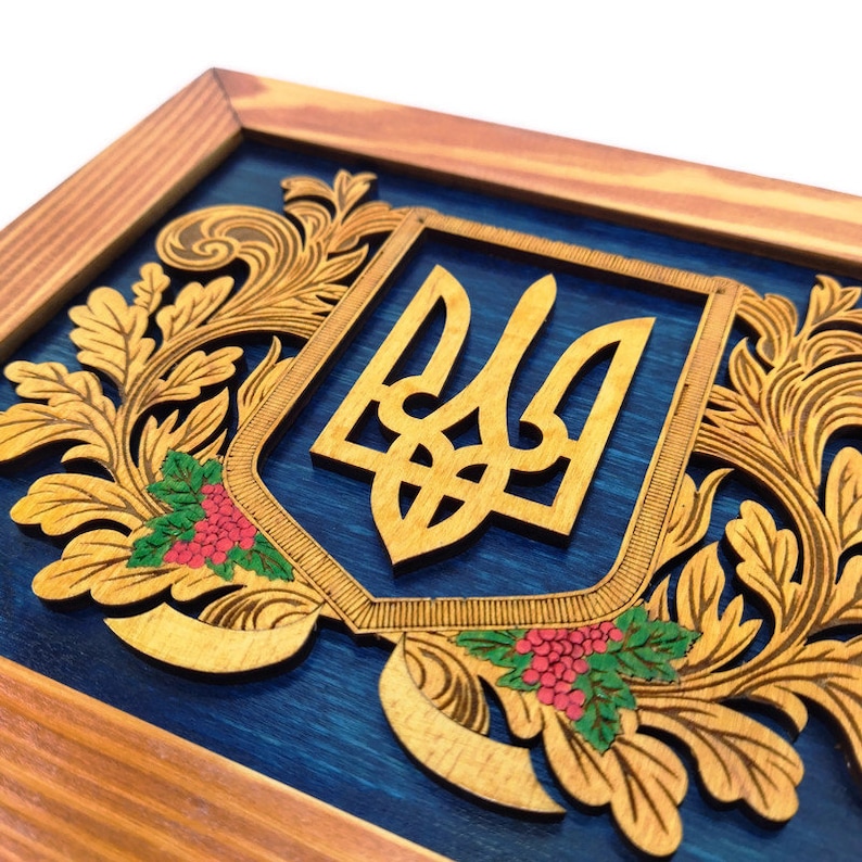 May include: A wooden plaque with a blue background and a carved wooden Ukrainian trident surrounded by floral designs. The trident is a symbol of Ukraine.