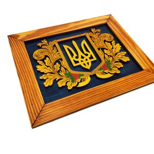 May include: A wooden framed wall art piece featuring a gold Ukrainian coat of arms with a blue background. The coat of arms is surrounded by gold floral designs.
